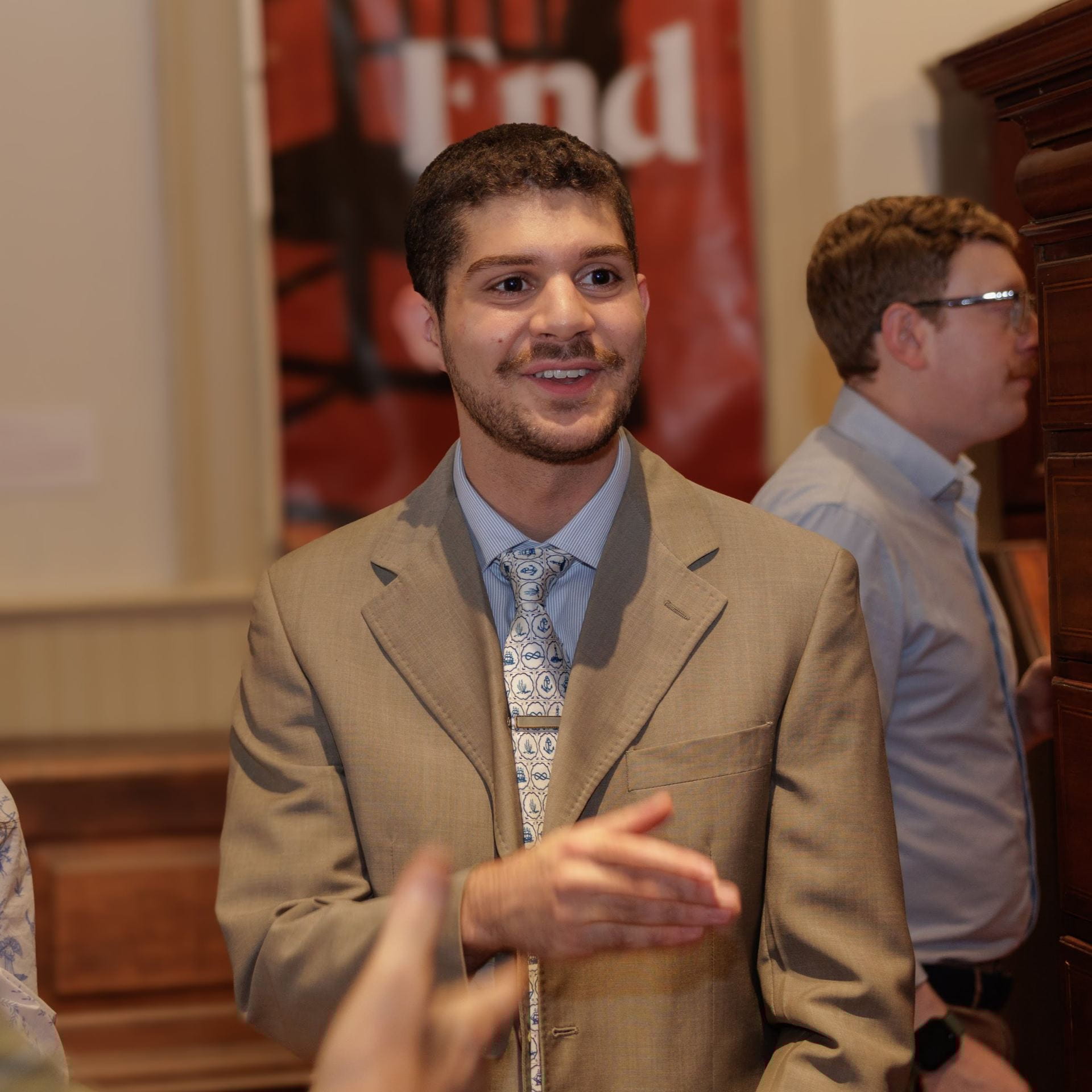 Half-length photo of a young person with dark hair and beard at an event, wearing a suit jacket and tie