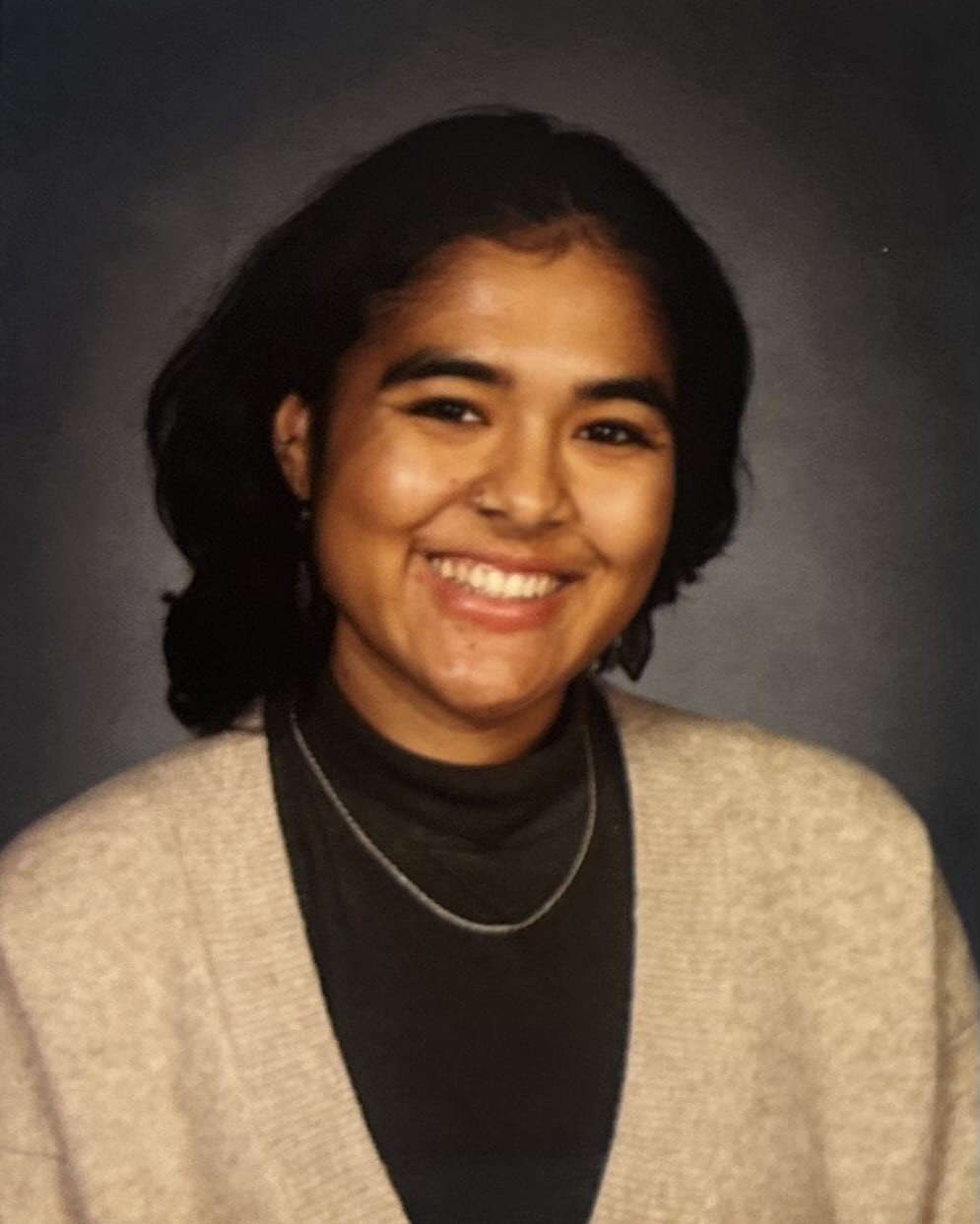 Portait photo of a young person in a black top and beige cardigan wearing a chain necklace