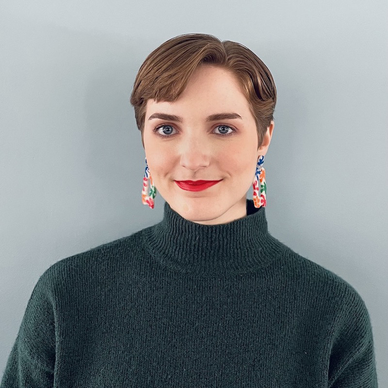 person with short brown hair, long dangly earrings, and black turtleneck smiling