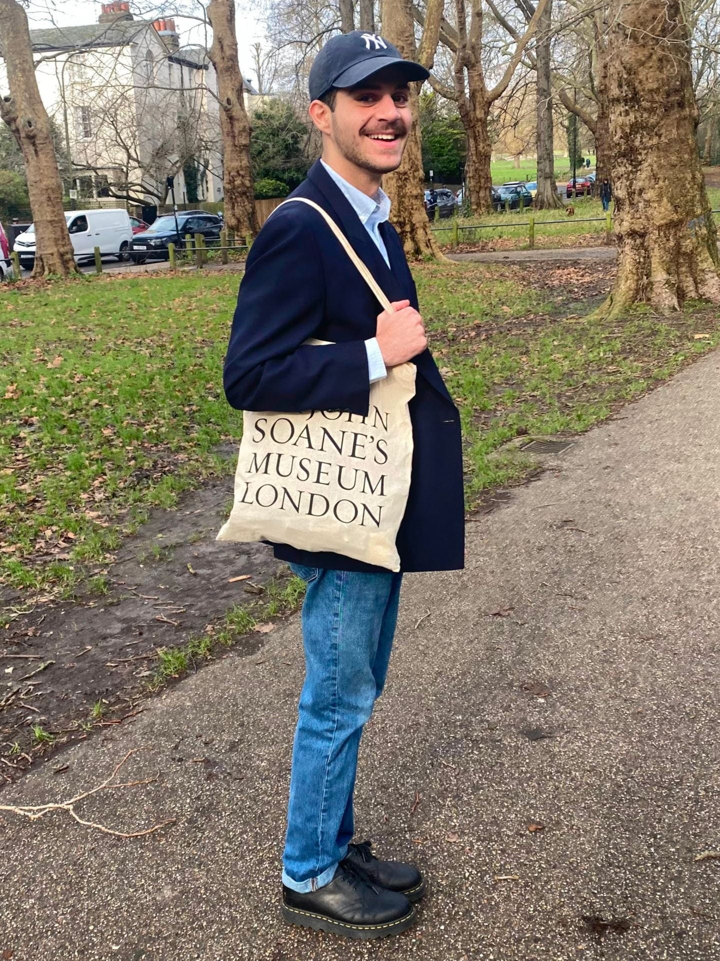 Full-length 3/4 view photo of a smiling young person in a jacket, jeans, cap holding a tote bag in a park.