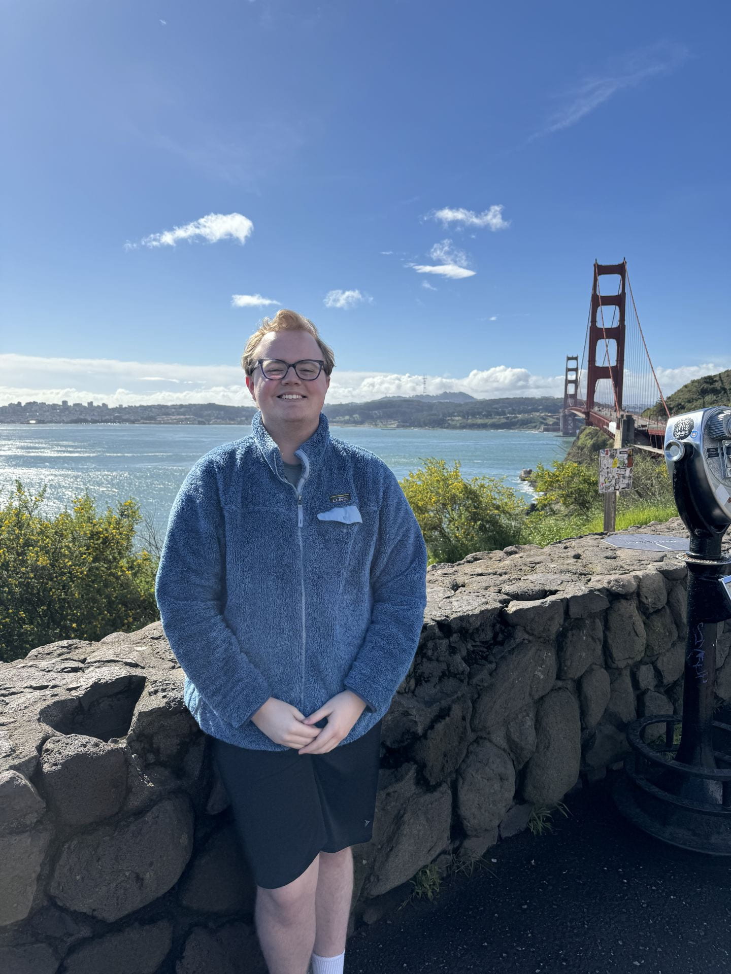 a young person in a blue fleece and shorts with glasses in front of the the Golden Gate Bridge in Sanfrancisco