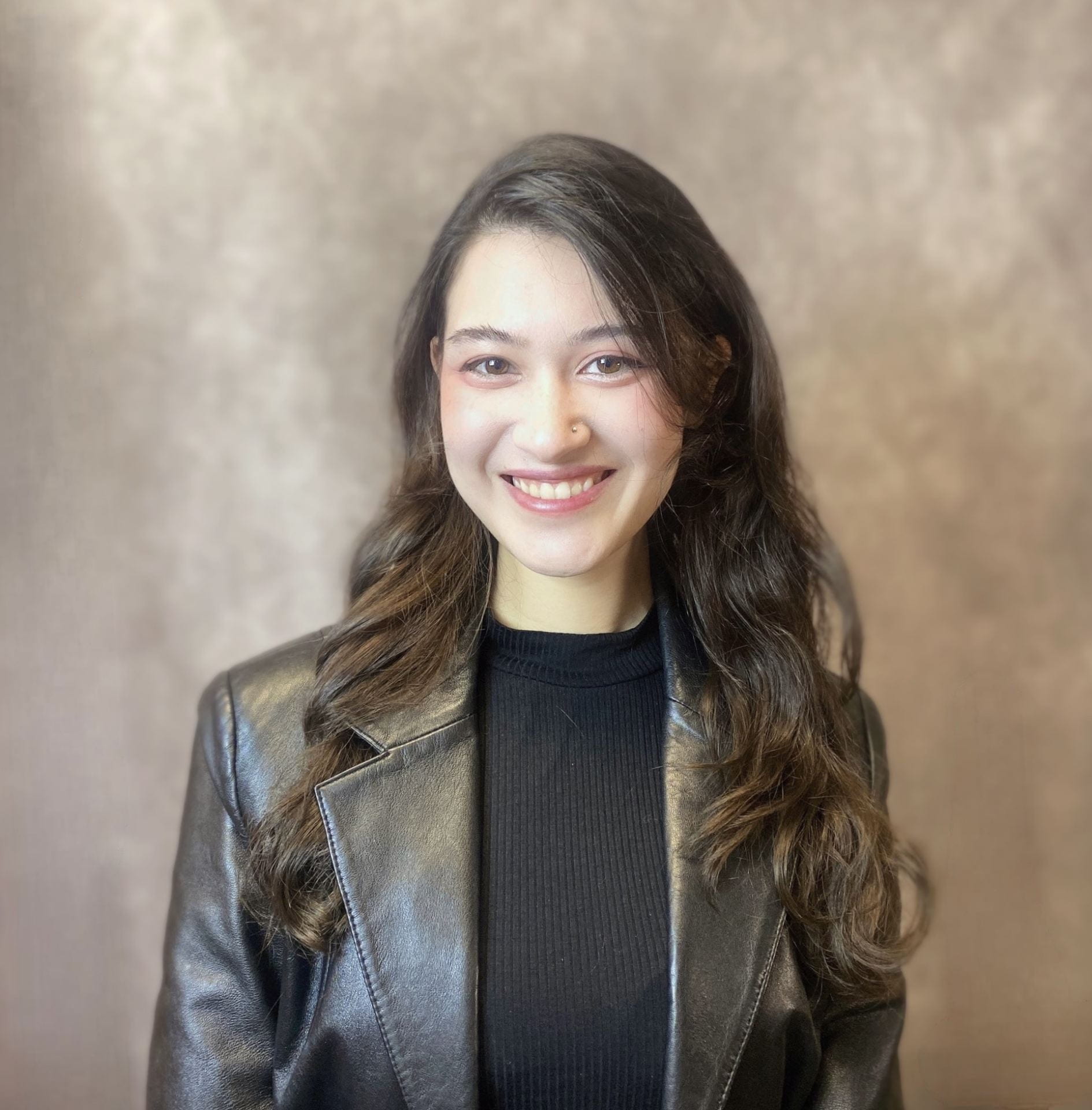 Portrait of a young person with long brown hair in a brown leather jacket and a brown shirt.
