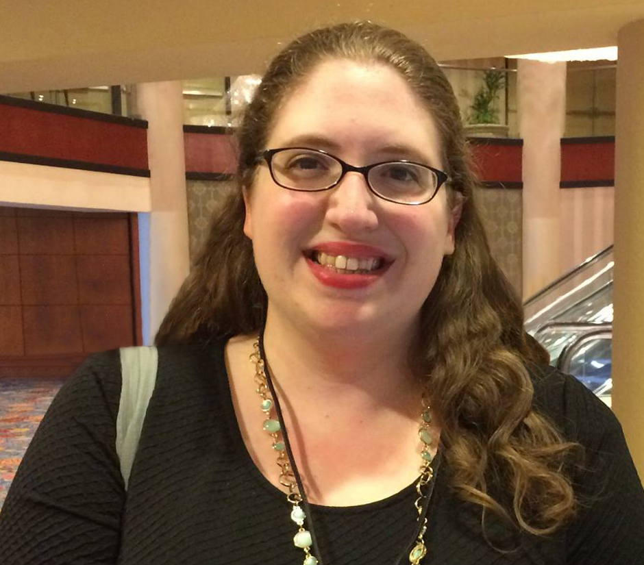 person with long brown hair, glasses, and a necklace smiling